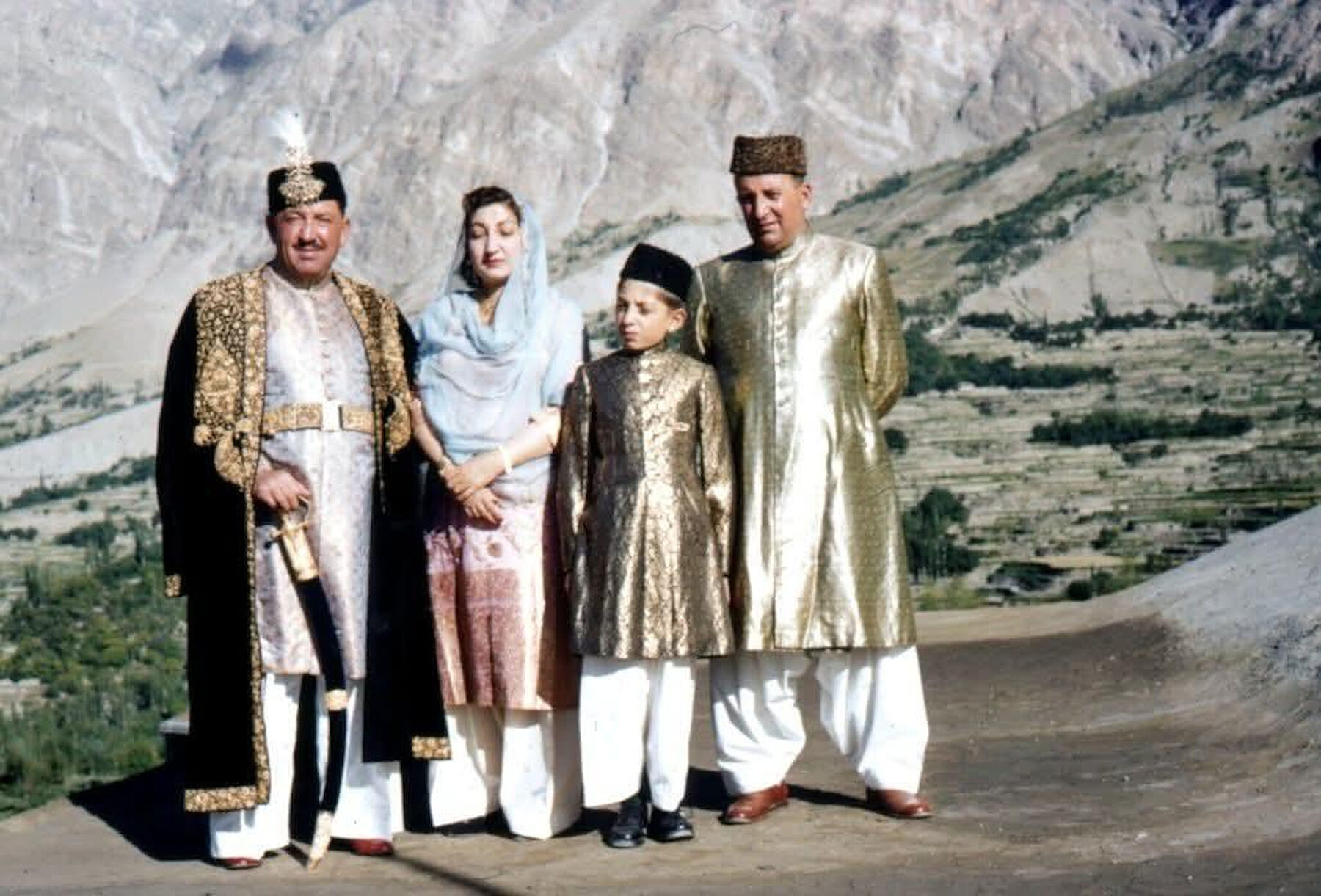 A family in gold ceremonial robes standing before the terraced slopes of the Hunza valley