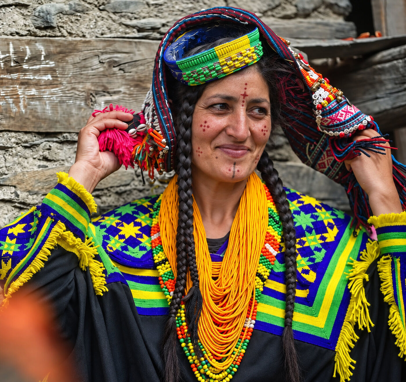 A Kalasha woman adjusting the pink woollen pom of her headdress against a weathered wooden wall