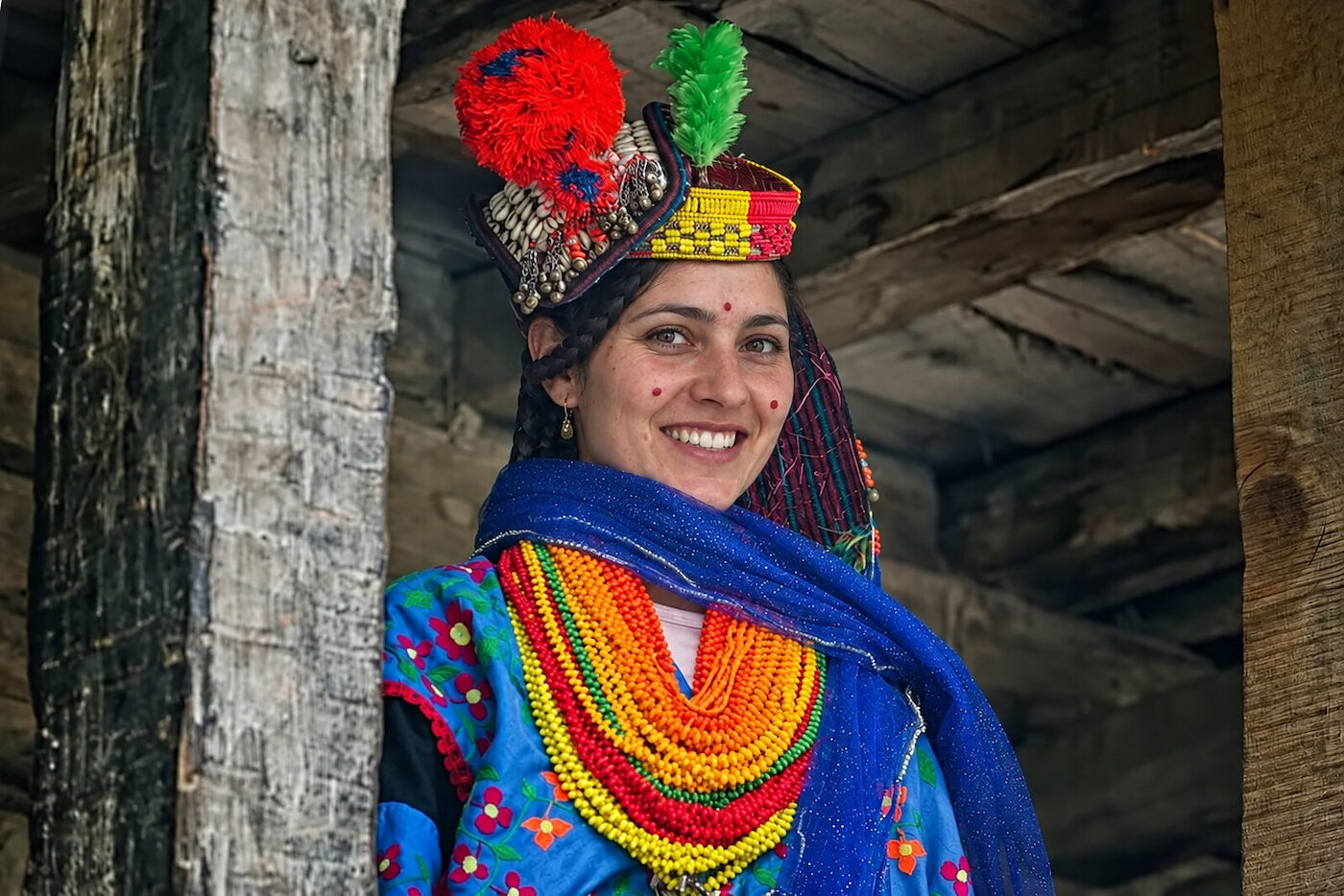 A Kalasha woman standing between timber posts in a red and green headdress and a blue shawl