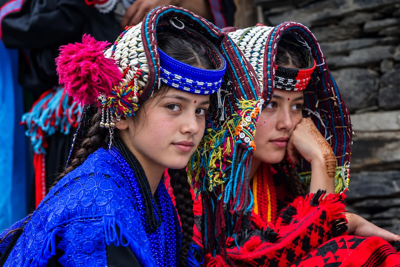 Two young Kalasha women seated together against a stone wall in embroidered dress